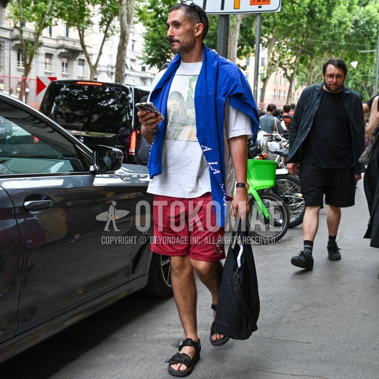 A summer men's outfit/coordination of plain black sunglasses, white graphic t-shirt, blue lettered long tee, red bottomed shorts, black sports sandals, and plain black briefcase/handbag.