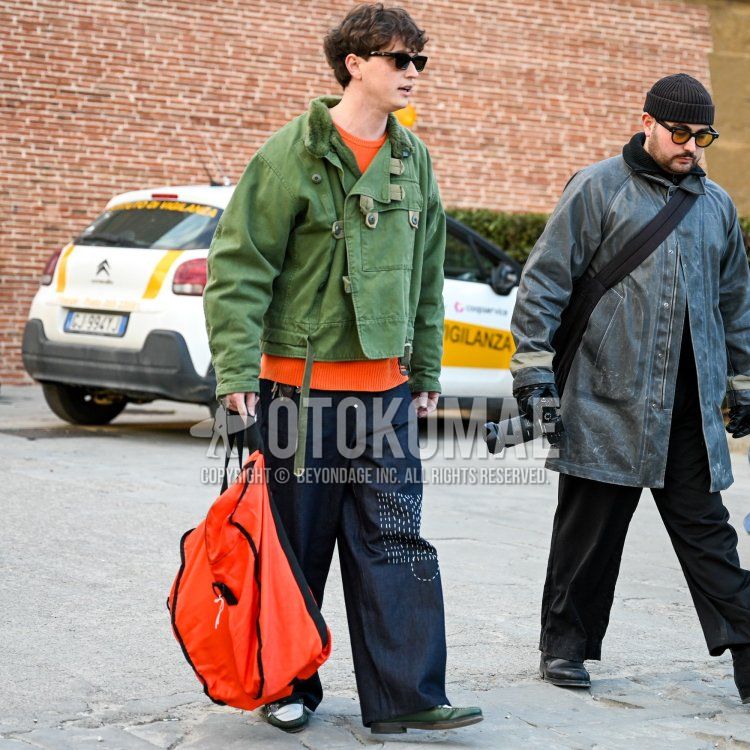 Men's fall/winter coordinate and outfit with solid black sunglasses, solid olive green outerwear, solid orange sweater, solid navy denim/jeans, green/white coin loafer leather shoes, and solid orange backpack.