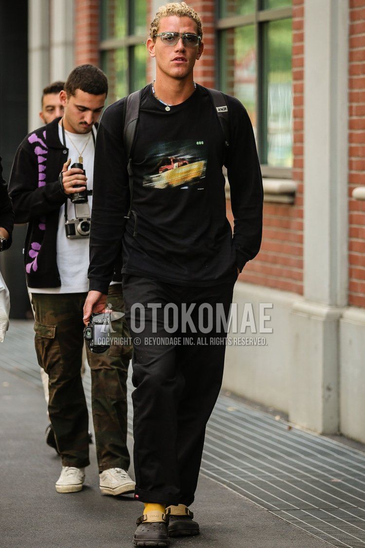 Men's spring/summer coordinate and outfit with plain silver glasses, black graphic long T, plain black denim/jeans, plain yellow socks, black sandals, and plain black backpack.