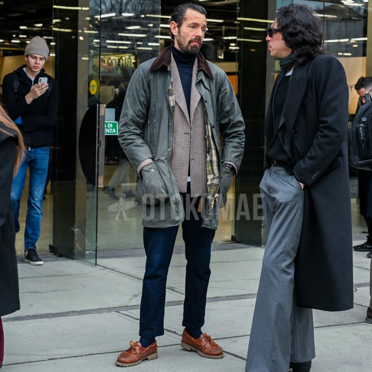 A man wearing a sage-colored waxed jacket that appears to be Barbour's BEAUFORT with center-pressed gray slacks and brown tasseled loafers. Holding a dark brown tote bag and coffee cup in his hand, he embodies the urban country look.