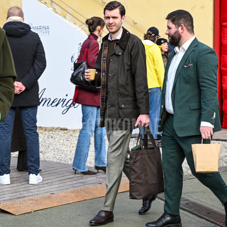 A man wearing what appears to be a "Beaufort" waxed jacket by Babur with gray slacks, tassel loafers, a tote bag and a cup of coffee in his hand