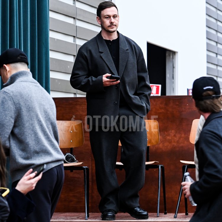 A man clad in a charcoal gray setup. A minimalist, urban mode style with a drop-shoulder jacket, wide pants, black innerwear, and voluminous black shoes on his feet.