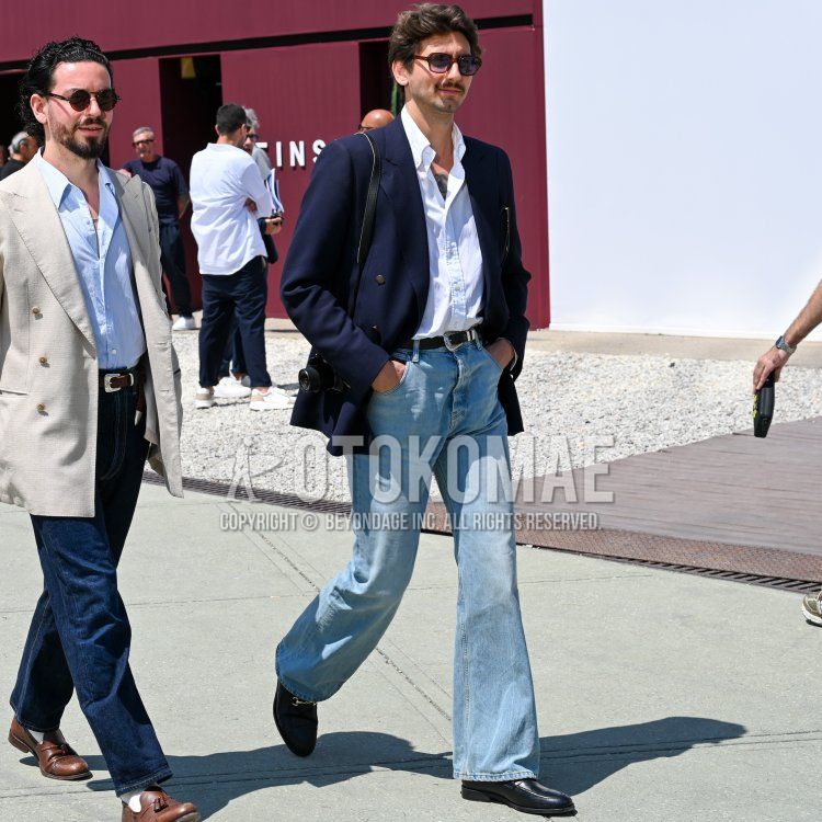 Men's spring/summer coordinate/outfit with brown tortoiseshell sunglasses, plain navy tailored jacket, plain white shirt, plain black leather belt, plain light blue denim/jeans, black bit loafer leather shoes, and plain black shoulder bag.