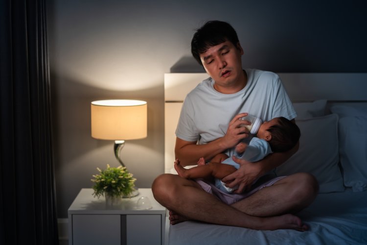 Sleepy and tired father feeding milk bottle to newborn baby on bed at night