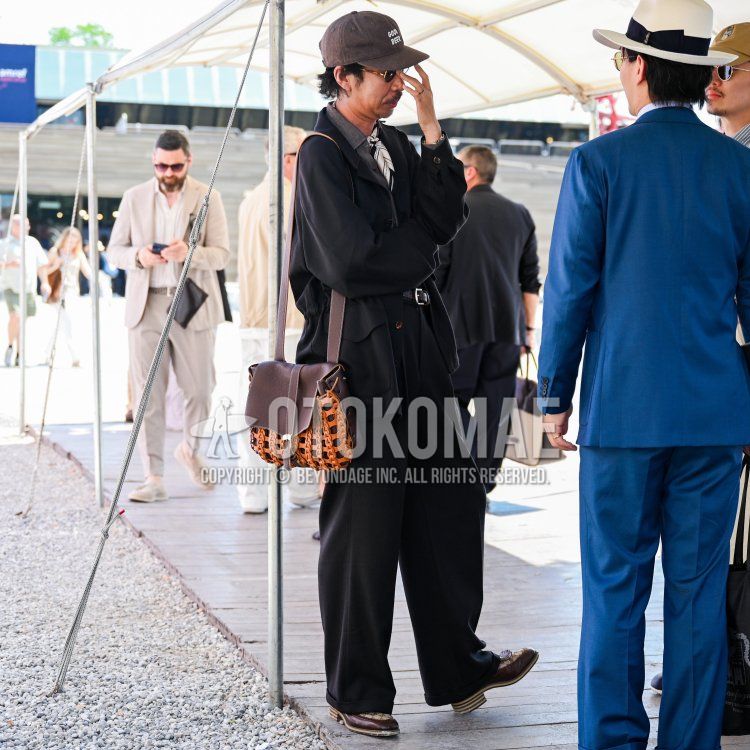 A spring/summer men's outfit featuring a brown-lettered baseball cap, plain gold sunglasses, a white bandana/neckchief, a plain black shirt jacket, a plain brown shirt, a plain black leather belt, plain black slacks, brown boots, and a plain orange-and-brown shoulder bag.