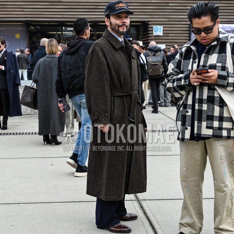 フィレンツェの街角、モダンな建物のファサードを背景に、30代後半から40代前半と見られる、引き締まった体格の男性が、短く整えられたダークブラウンの髪と、手入れの行き届いたフルビアードをたくわえ、ブラックの「L'ART DE L'AUTOMOBILE」ロゴ入りベースボールキャップを被り、その下にはライトブルーのストライプシャツにパープルのネクタイを締め、ダークブラウンのヘリンボーン柄の厚手のベルテッドロングコートを羽織り、ネイビーのワイドスラックスとダークブラウンのレザーキャップトゥダービーを合わせ、右手薬指にはゴールドのリングを着用した、クラシックとカジュアルが融合した洗練されたスタイルで佇んでいる。
