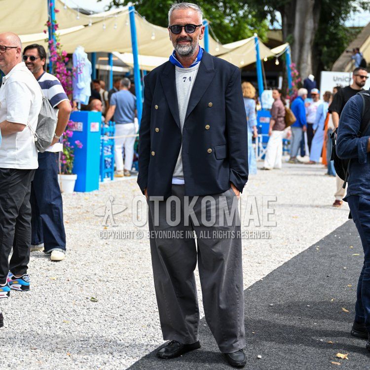 A men's spring/summer outfit featuring plain black sunglasses from Boston, a blue stole scarf, a plain black tailored jacket, a white T-shirt with lettering, plain gray denim jeans, plain gray pleated pants, and black leather loafers.