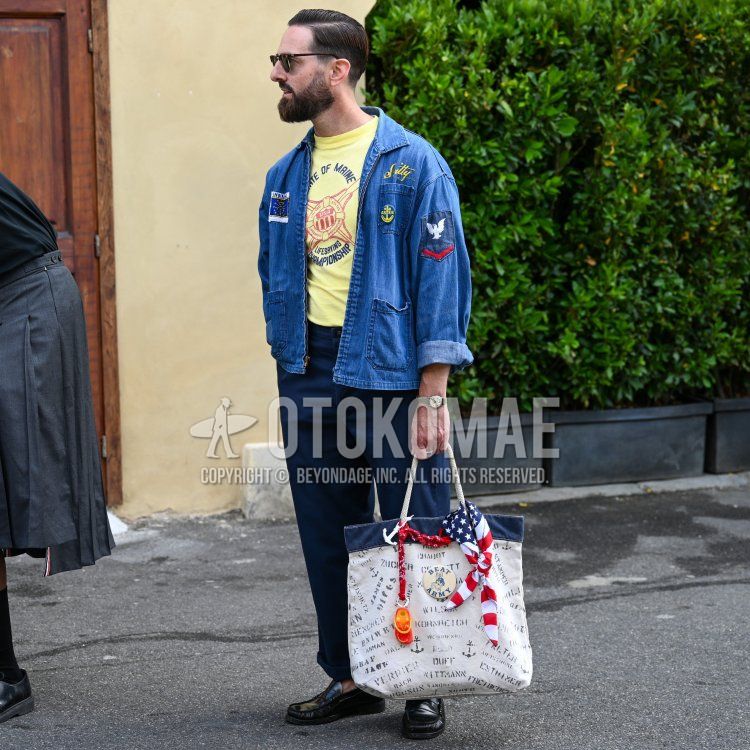 A spring/summer men's outfit featuring plain black sunglasses, a multicolored scarf, a denim jacket with a blue accent, a T-shirt with yellow lettering, plain navy slacks, black leather coin loafers, and a tote bag with white lettering.