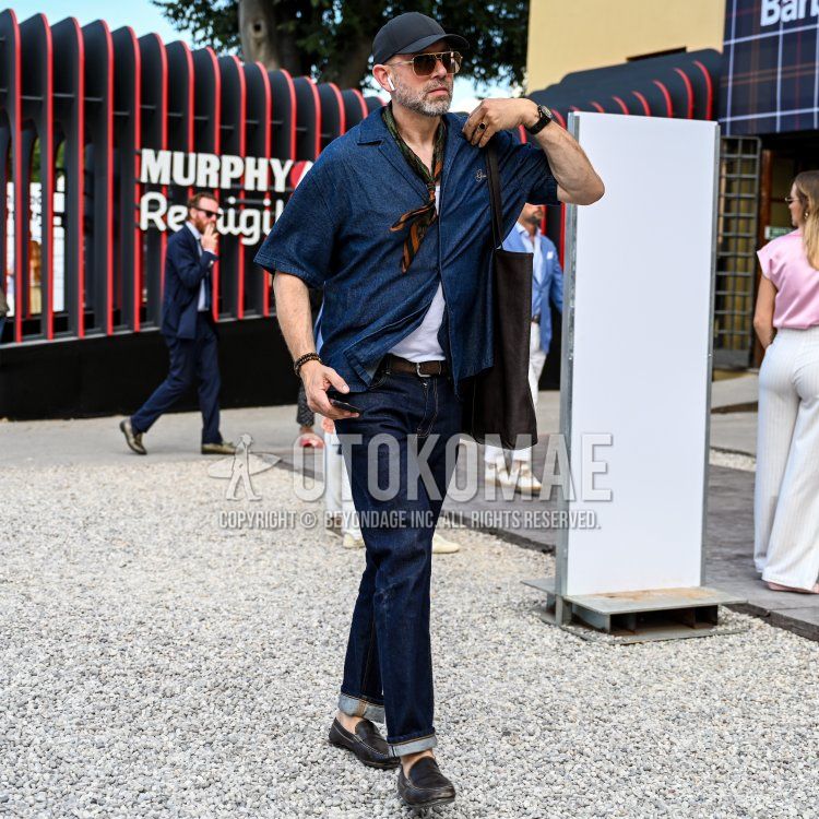 At an outdoor venue in Florence, a man who appears to be in his 40s or 50s walks across a ground paved with white pebbles. He is distinguished by his closely cropped hair and a well-groomed salt-and-pepper beard.He is wearing a black baseball cap and gold-framed aviator sunglasses, with a white wireless earbud in his left ear.Around his neck, he wears a brown, orange, green, and black paisley-patterned silk scarf; over a white crew-neck T-shirt, he wears a dark blue short-sleeved denim shirt; and he pairs this with dark indigo straight-leg jeans, rolled up at the ankles and cinched at the waist with a dark brown leather belt.On his feet are dark brown leather loafers; he carries a dark brown leather tote bag over his left shoulder; on his left wrist is a watch with a black strap; on his right wrist is a dark brown beaded bracelet; on his right ring finger is a ring with a black stone; and he is holding a smartphone in his right hand.