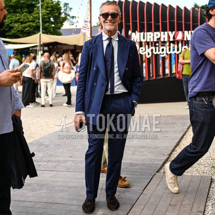 フィレンツェの晴れ渡った屋外イベント会場で、木製の通路と砂利の地面を背景に、50代後半から60代前半と見られる、引き締まった体型の男性が、短く整えられたグレーの髪と髭をたくわえ、笑顔でカメラを見つめている。彼はネイビーの細いストライプが入ったダブルブレストスーツを着用し、その下にはライトブルーのストライプシャツとネイビーのレギュラータイを合わせている。足元は、トンボのような刺繍が施されたダークブラウンのスエードスリッポンローファーで、左手にはスマートフォンを持ち、右手はスーツのポケットに入れている。顔にはウェリントン型の黒いサングラスをかけ、左手首にはシルバーの腕時計が光る、洗練されたイタリアンクラシックスタイル。