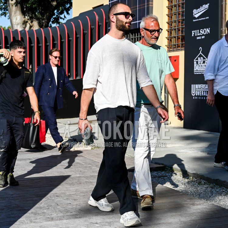 On a sunny day in Florence, against a backdrop of wooden boardwalks and buildings featuring black and red accents, a man in his thirties—with closely cropped hair and a well-groomed, thick beard—with a deep tan, wearing black square-frame sunglasses, a loose-fitting white linen knit T-shirt, and navy pleated wide-leg pants,paired with white and gray chunky sneakers; a slim silver bracelet on his left wrist; and a small camera in his right hand as he walks lightly.