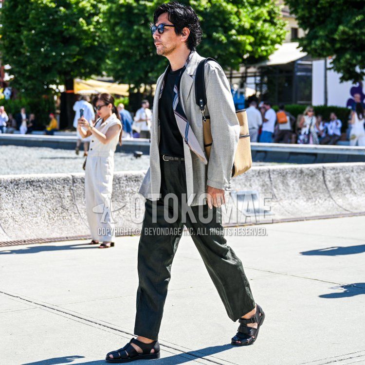 On a sunny day in Florence, against a backdrop of lush greenery, a man who appeared to be in his late 30s to early 40s—with slightly tanned skin, wavy black hair swept back casually, and a neatly trimmed beard—wore a light gray linen-blend jacket,wearing a black crew-neck T-shirt underneath, paired with dark green relaxed-fit trousers and black leather fisherman sandals on his feet.He wore a navy and beige striped scarf around his neck, carried a beige canvas tote bag over his right shoulder, and sported black sunglasses and a black leather belt as he walked along in his light, summery outfit.
