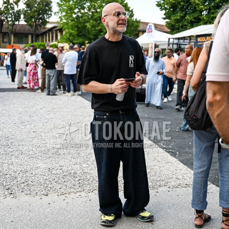 フィレンツェの屋外イベント会場の石畳の広場で、40代後半から50代前半くらいでややがっしりとした体型の男性が、坊主頭に白髪交じりの短く整えられた髭をたくわえ、やや日焼けした肌で佇んでいる。彼は、フロントに「R END.」のようなロゴがプリントされた黒のオーバーサイズTシャツに、濃紺のワイドデニムパンツ、足元には蛍光イエローと黒の切り替えが特徴的なスニーカーを合わせている。顔にはシルバーのツーブリッジフリップアップ式サングラスをかけ、左手首にはシルバーのバングル、右手首には黒いブレスレットを着用し、黒いレザーベルトを締め、右手にスマートフォンとペットボトルを持っている、モダンでリラックス感のあるストリートカジュアルスタイル。