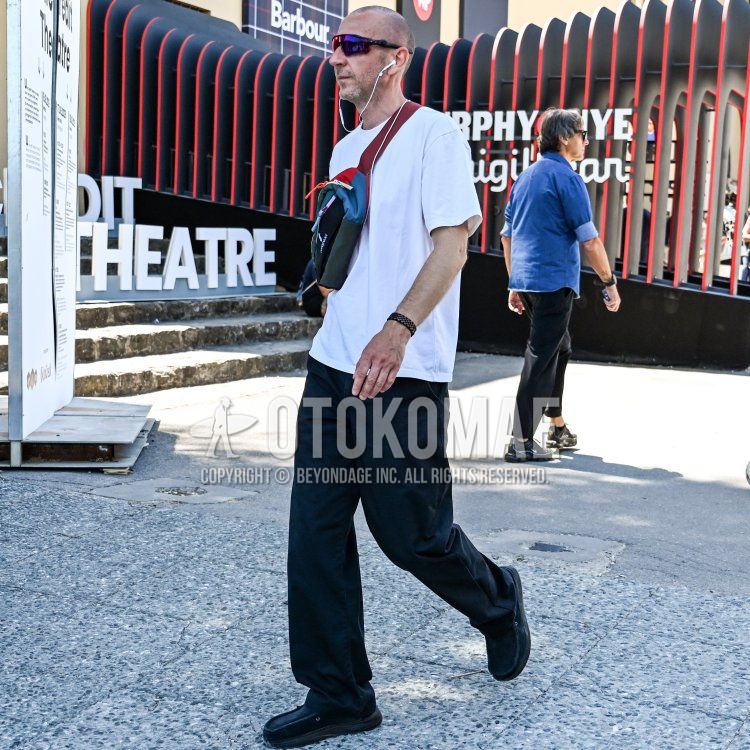 フィレンツェの晴れた日、黒と赤のストライプのモダンな建物の前で、短く刈り込んだ髪型で顔に薄い髭の跡がある、引き締まった体型の40代後半から50代前半の男性が、白いクルーネックの半袖Tシャツに黒のワイドレッグパンツを合わせ、足元には黒の厚底カジュアルシューズを履き、右肩からカラフルなクロスボディスリングバッグを斜めがけし、左手首にはダークカラーのビーズブレスレットを着用。さらに、パープル系のミラーレンズと黒いフレームのスポーティーなラップアラウンドサングラスをかけ、白い有線イヤホンを装着して歩いている姿。