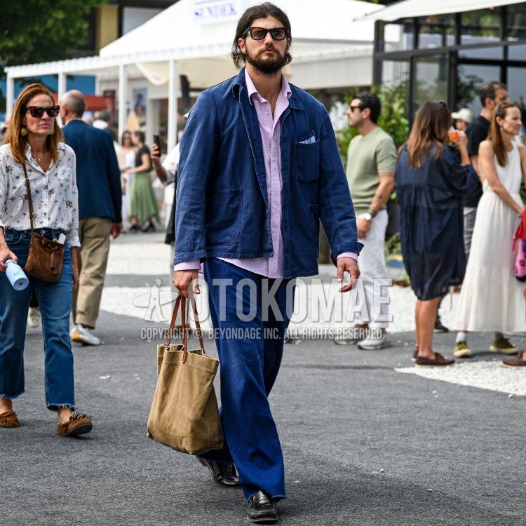 フィレンツェの屋外会場で、白いテントや木々を背景に、ダークブラウンのミディアムロングのウェーブヘアに整えられたフルビアードと口髭をたくわえ、日焼けした肌の20代後半から30代前半の男性が、ブラックのウェリントン型サングラスをかけ、ネイビーのワークジャケットを羽織り、その下にはピンクと白のギンガムチェックの長袖シャツを襟元を開けて着用し、ダークブルーのワイドレッグパンツとブラックのペニーローファーを合わせ、右手にはベージュのキャンバストートバッグを持って、リラックスしたスマートカジュアルな装いで歩いている。