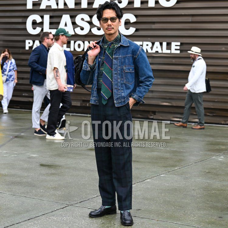 A spring/summer men's outfit featuring clear brown solid-color sunglasses, a solid blue denim jacket, a green checkered shirt, green and navy checkered slacks, solid green socks, black leather coin loafers, a solid black leather shoulder bag, and a green and navy regimental tie.