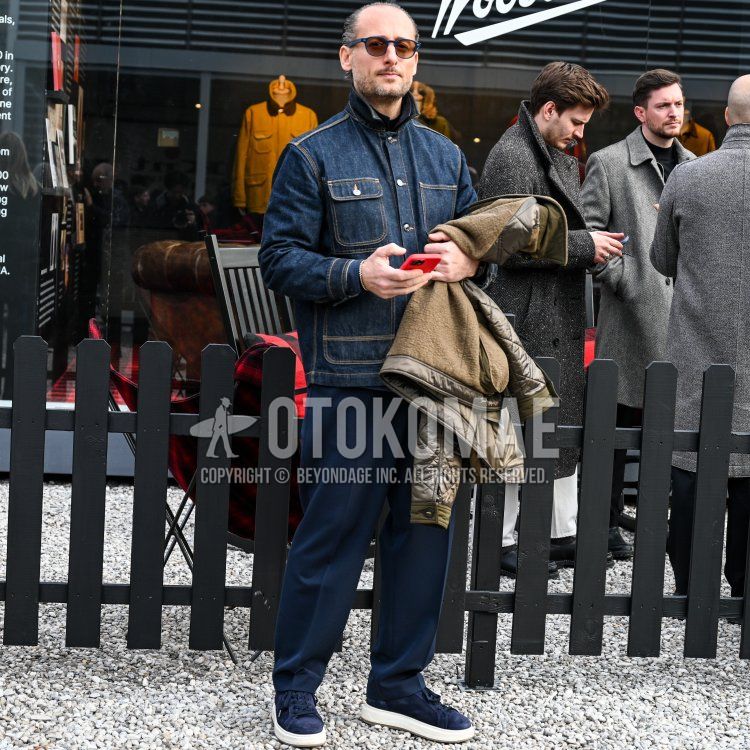 On the cobblestone streets of Florence, against a backdrop of glass-fronted shops and black wooden railings, stood a man who appeared to be in his late 30s to early 40s, with a toned physique and dark brown hair slicked back,and a neatly trimmed goatee, stands in a sophisticated, casual style. He wears navy round-frame sunglasses and is dressed in a dark blue denim jacket paired with a black mock-neck top, navy relaxed-fit trousers, and navy suede sneakers.He holds a red smartphone in his right hand and is carrying an olive-green quilted jacket in his left arm.