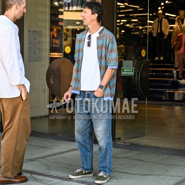 フィレンツェの店舗のガラス扉の前で、40代後半から50代前半に見える、平均的な体格で、ダークブラウンの短くカールした髪をやや無造作にセットし、薄い髭をたくわえ、やや日焼けした肌の男性が、白いクルーネックTシャツの上にティール、パープル、オリーブグリーンの幾何学模様が特徴的な半袖ニットカーディガンを羽織り、ライトブルーのウォッシュ加工されたリラックスフィットジーンズ、足元にはオリーブグリーンのキャンバススニーカーを着用。首元には細いシルバーチェーンのネックレス、左手首にはスクエア型のデジタルウォッチ、右手首にはビーズブレスレットをつけ、サングラスをTシャツの襟元にかけ、右手にはタバコを持ちながら、左側に立つ人物と会話している。