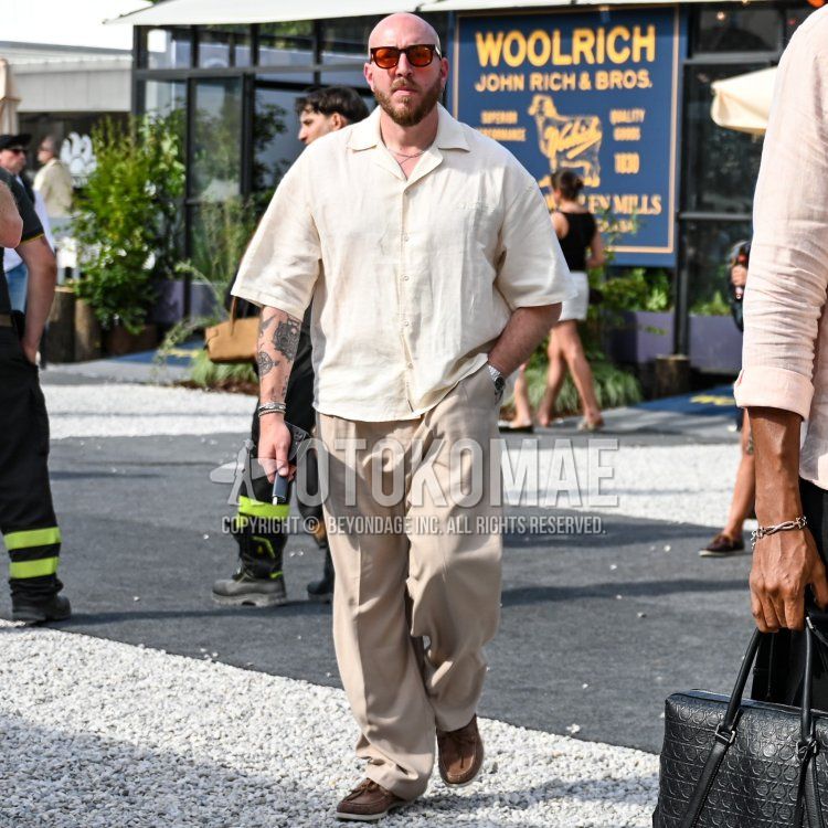 フィレンツェの屋外会場、晴れた日差しのもと、「WOOLRICH」の看板を背景に、がっしりとした体格でスキンヘッド、整えられたゴートヒゲをたくわえ、こんがりと日焼けした肌の30代後半から40代前半の男性が、オフホワイトのオーバーサイズリネン開襟シャツにベージュのワイドスラックス、足元にはブラウンのスエードデッキシューズを合わせた、リラックス感のあるサマースタイルで佇んでいる。彼はオレンジレンズのスクエア型サングラスをかけ、首元にはシルバーのチェーンネックレス、左耳にはフープピアス、左手首には複数のシルバーブレスレットとメタルバンドの腕時計を身につけ、右手には黒い型押しレザーのブリーフケースを持っている。