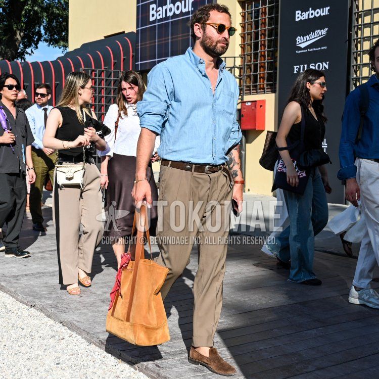 フィレンツェの晴れ渡った日、Barbourのロゴが見える建物の前、木製の通路を歩く、30代後半から40代前半で中肉中背、短くカールしたダークブラウンの髪と整えられたフルビアード、健康的に日焼けした肌の男性が、左腕にタトゥーを覗かせながら、ライトブルーのシャンブレーシャツの袖を捲り、プリーツ入りのカーキ色チノパンを合わせ、足元にはダークブラウンスエードのローファーを履き、ダークブラウンのレザーベルト、アンバーフレームのラウンド型サングラス、左手首にはゴールドの細身のブレスレット、右手首にはダークブラウンのレザーブレスレットを着用し、左手には赤いスカーフが結ばれたキャメル色のスエードトートバッグを持ち、右手にはスマートフォンを持って佇む姿。