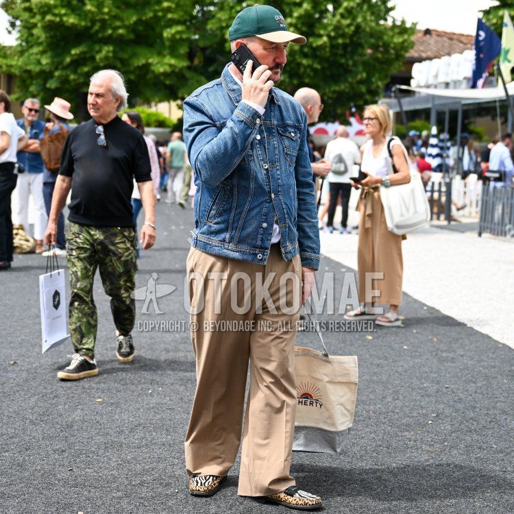 A spring/summer men's outfit featuring a green-lettered baseball cap, a plain blue denim jacket, a plain white shirt, plain beige slacks, plain beige pleated pants, multicolored leather coin loafers, and a tote bag with a white accent.